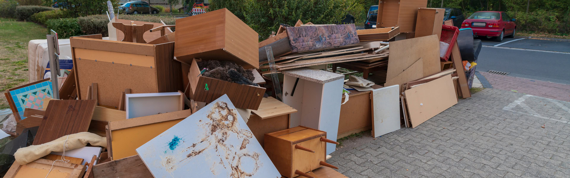 Ein Stapel alter Holzmöbel liegt nahe einem Parkplatz auf dem Gehweg im Freien.