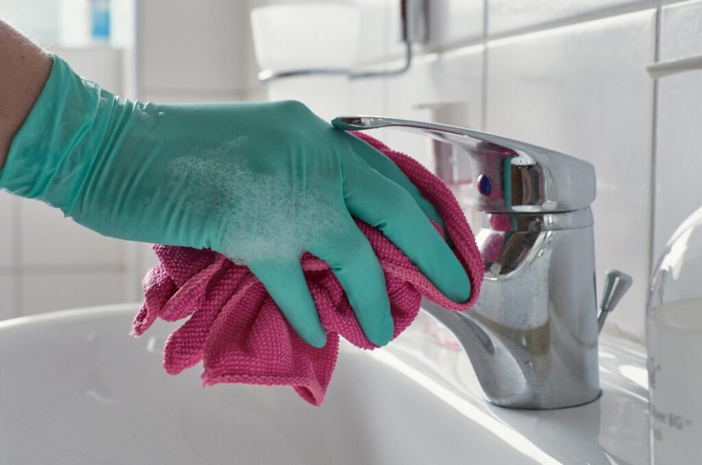Hand mit Latexhandschuhen und Reinigungstuch reinigt Wasserhahn des Waschbeckens